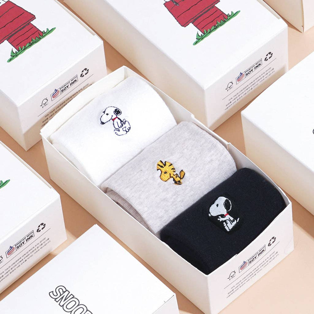 Three pairs of socks with cartoon Snoopy characters in a box, surrounded by more boxes. The perfect gift for snoopy lovers